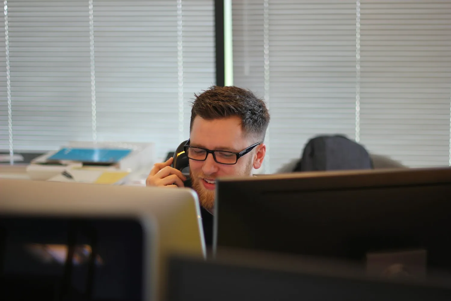 Un homme à lunettes sourit au téléphone derrière ses écrans, dans un bureau aux stores baissés