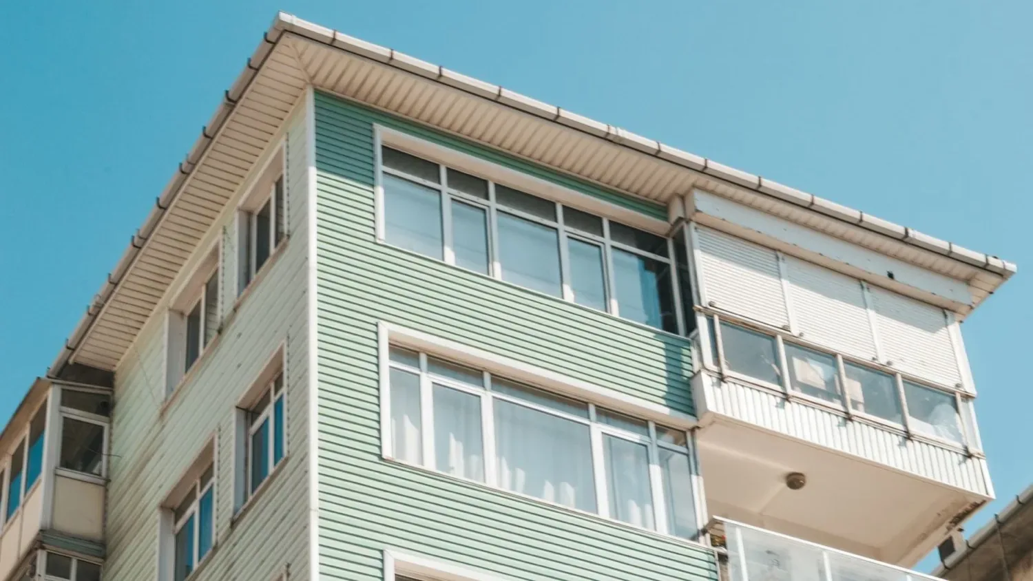 Haut d’un immeuble d’habitation au bardage vert d’eau, fenêtres blanches et balcon fermé, sous un ciel bleu