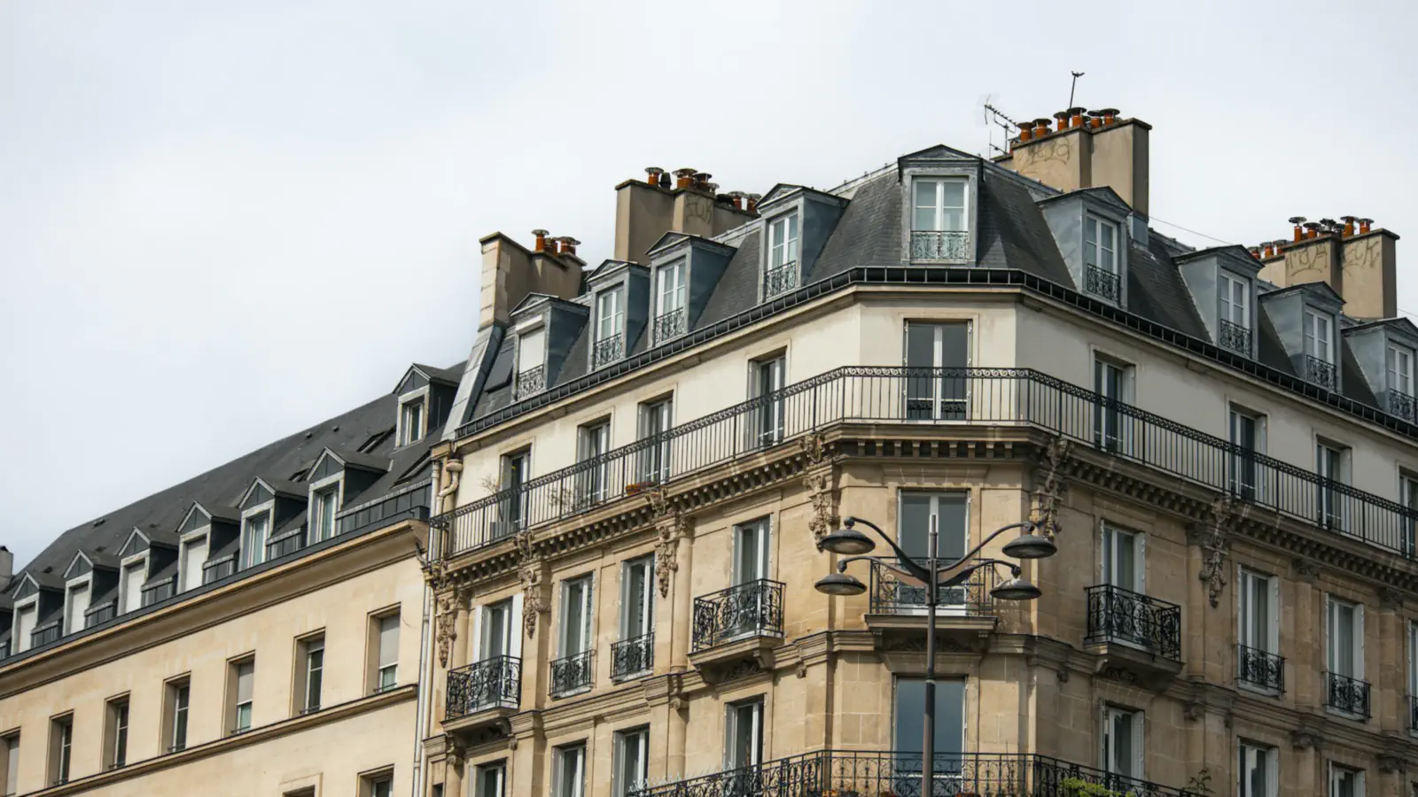 Immeuble haussmannien d’angle en pierre de taille : toit mansardé à lucarnes, cheminées, balcon filant en fer forgé et lampadaire, sous un ciel blanc