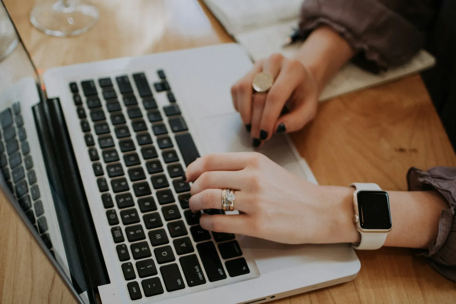 Des mains baguées tapent sur le clavier d’un ordinateur portable posé sur une table en bois, une montre connectée au poignet