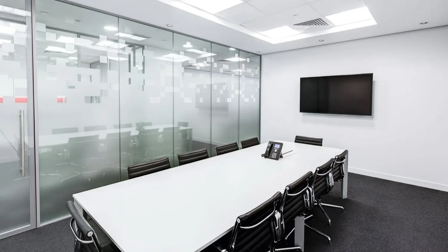 Salle de réunion vitrée et vide, table blanche, fauteuils noirs et écran mural éteint