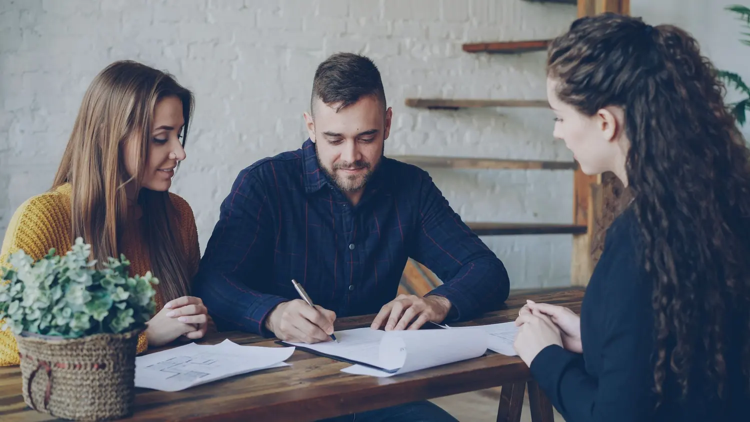 Un homme signe un document à une table en bois, une femme à ses côtés et une autre en face de lui