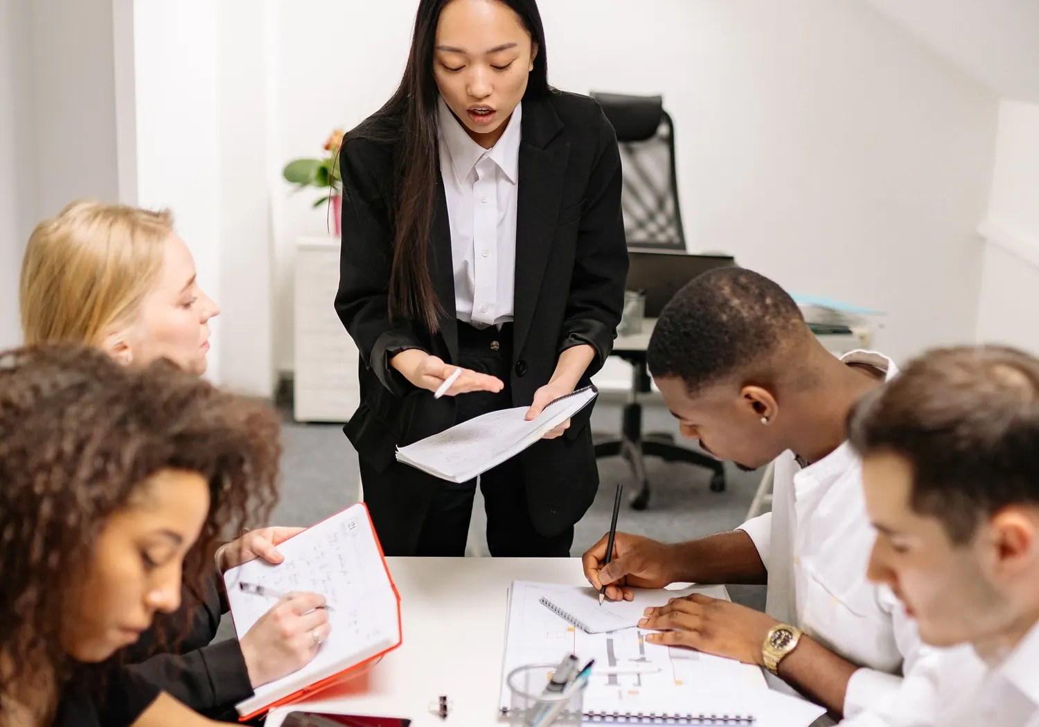 Une responsable debout, un document à la main, face à son équipe assise qui prend des notes