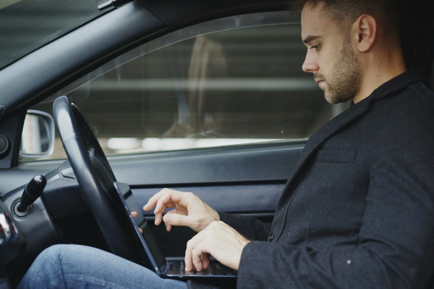 Un négociateur en veste travaille sur son ordinateur portable, assis au volant de sa voiture entre deux rendez-vous