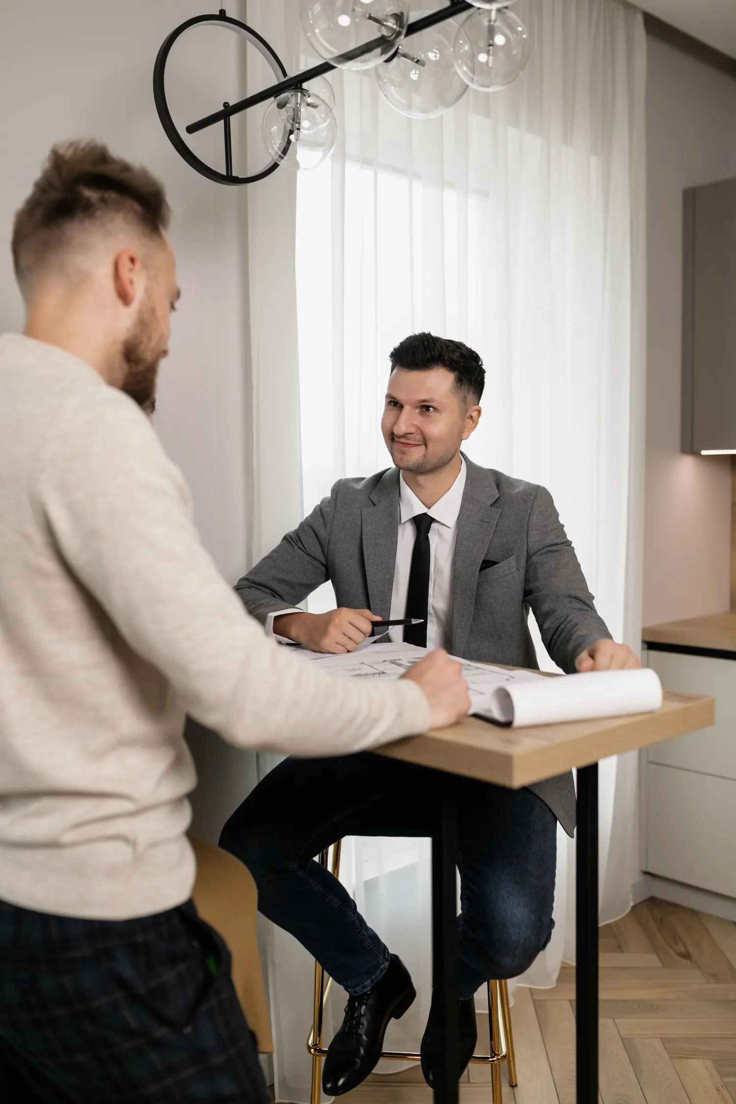 Un conseiller en veste grise échange avec un client autour de plans posés sur une table haute